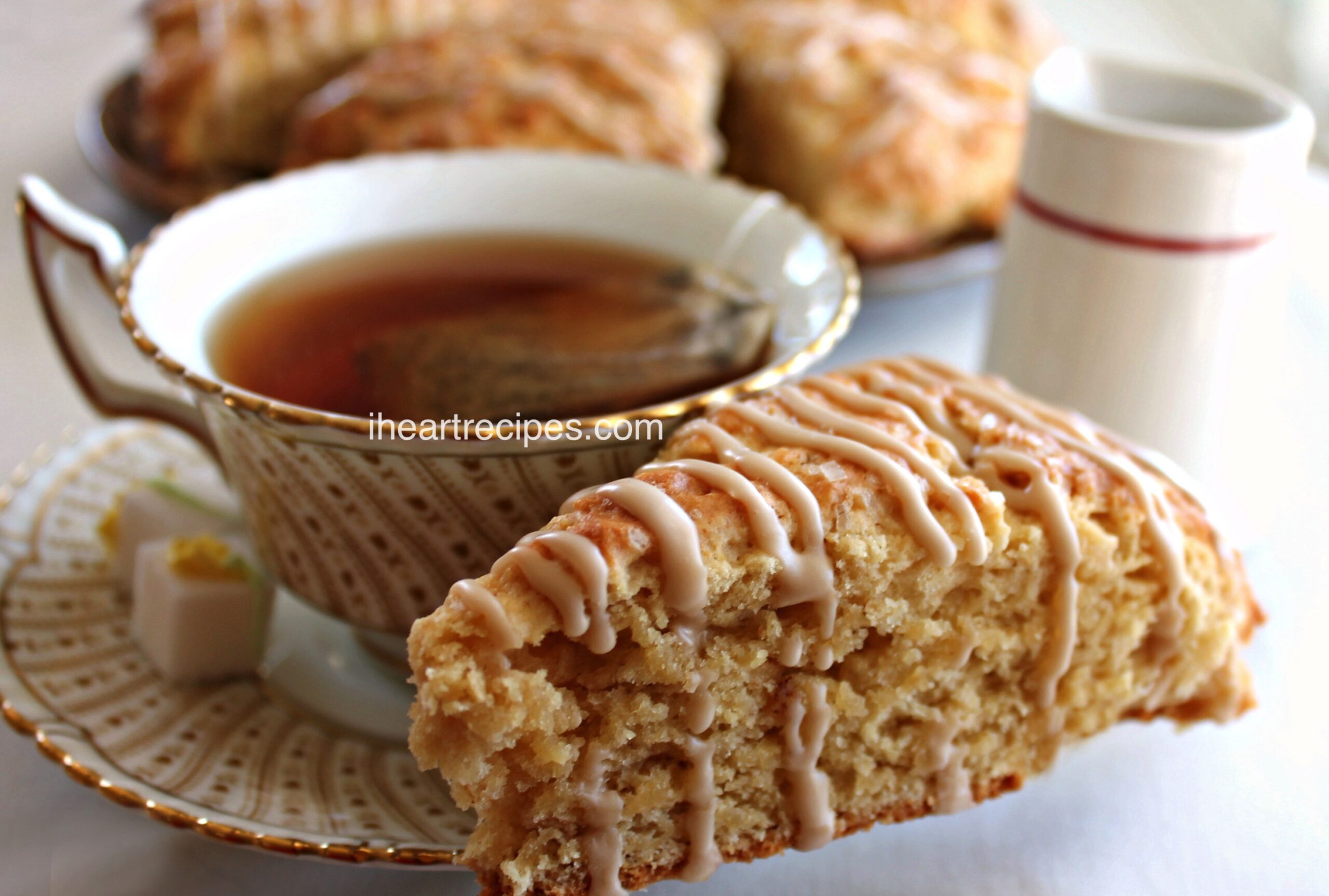 A homemade brown butter maple scone with maple icing sits on a tea saucer with a cup of warmed, freshly brewed tea,