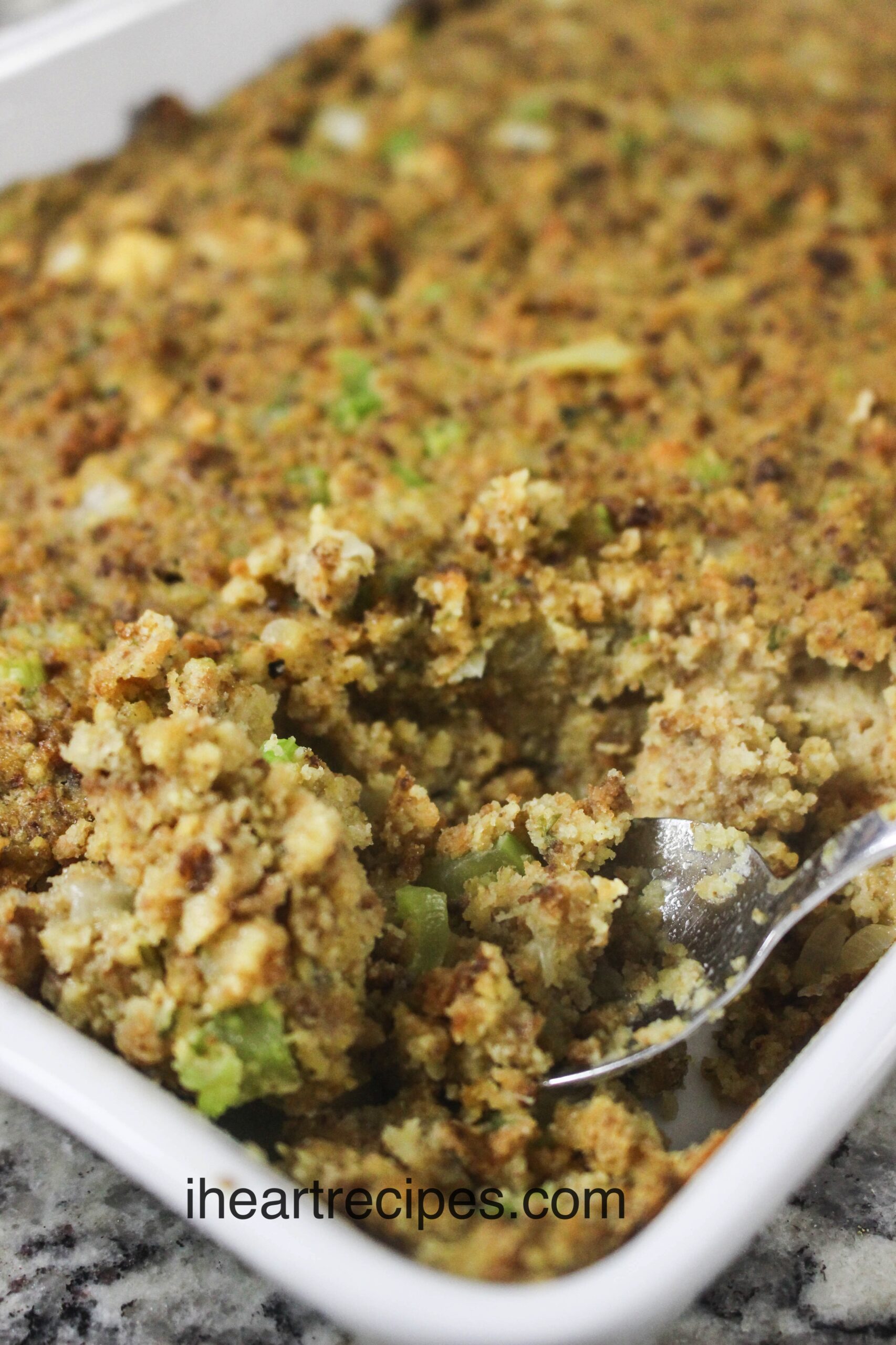 A large casserole dish of homemade cornbread dressing. A spoon scoops a serving from the corner, showing the dressing's moist texture and chunks of celery and onions throughout the dressing.