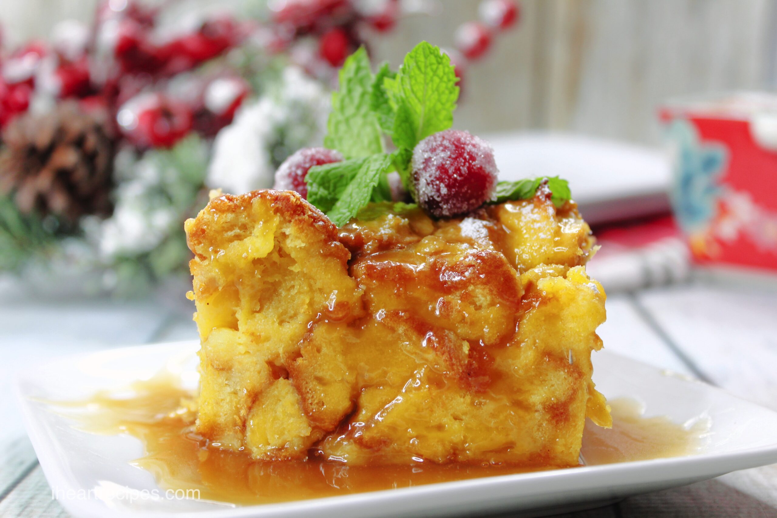 A perfect slice of moist bread pudding doused with maple syrup, served on a plate and garnished with fresh raspberries, mint leaves, and a dusting of powdered sugar.