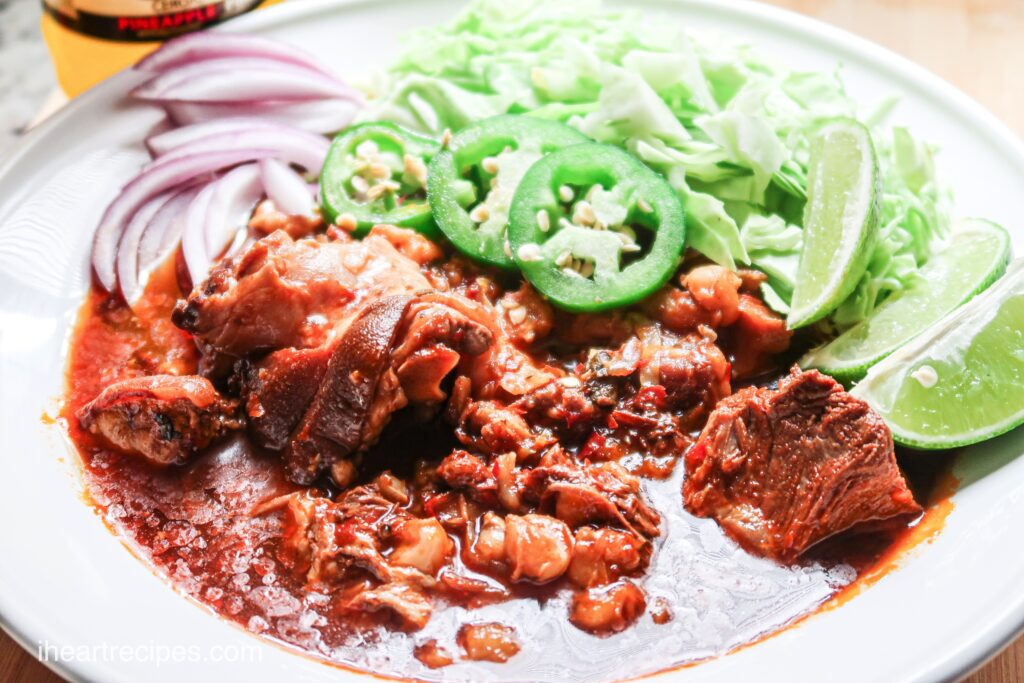 A flavorful slow cooker pork pozole stew served with lime, jalapeno, and red onion slices and fresh cabbage assembled in a white bowl.