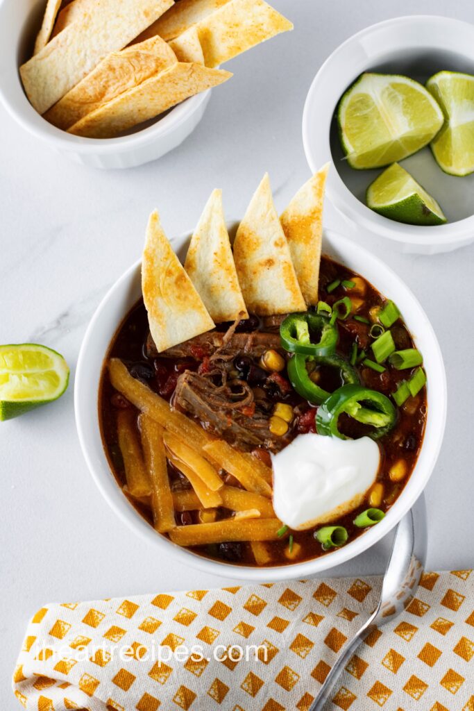 A white bowl filled with beef and veggies in a warm, savory broth garnished with pita chips.