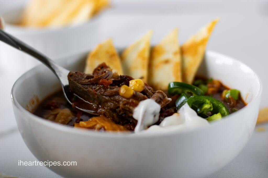 A silver spoon lifts a tender piece of brisket out of a white bowl filled with chili. 