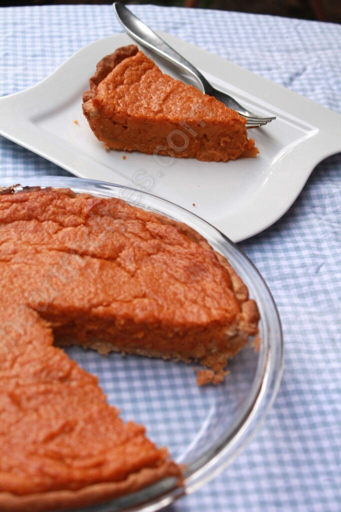 A whole sweet potato pie in a glass pie dish with one slice removed sits in the forefront of the image. In the background, a single slice of pie is served on a white square plate with a fork. The pie is set out on a blue and white gingham-patterned tablecloth.