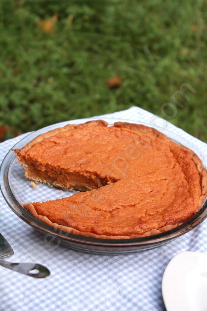 A whole sweet potato pie with one slice missing sits in a glass pie dish on the corner of a blue and white gingham tablecloth.