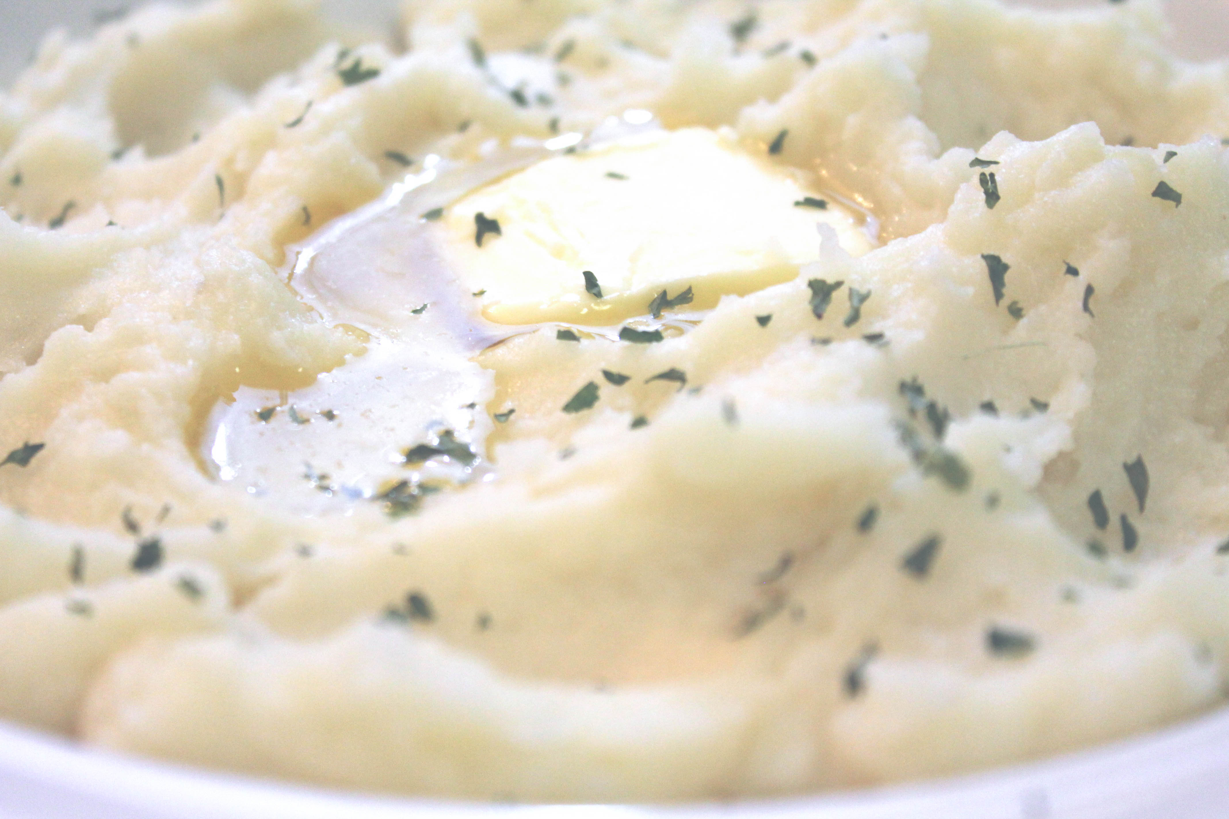 A close up image of creamy homemade mashed potatoes, garnished with parsley flakes and a pat of melting butter.