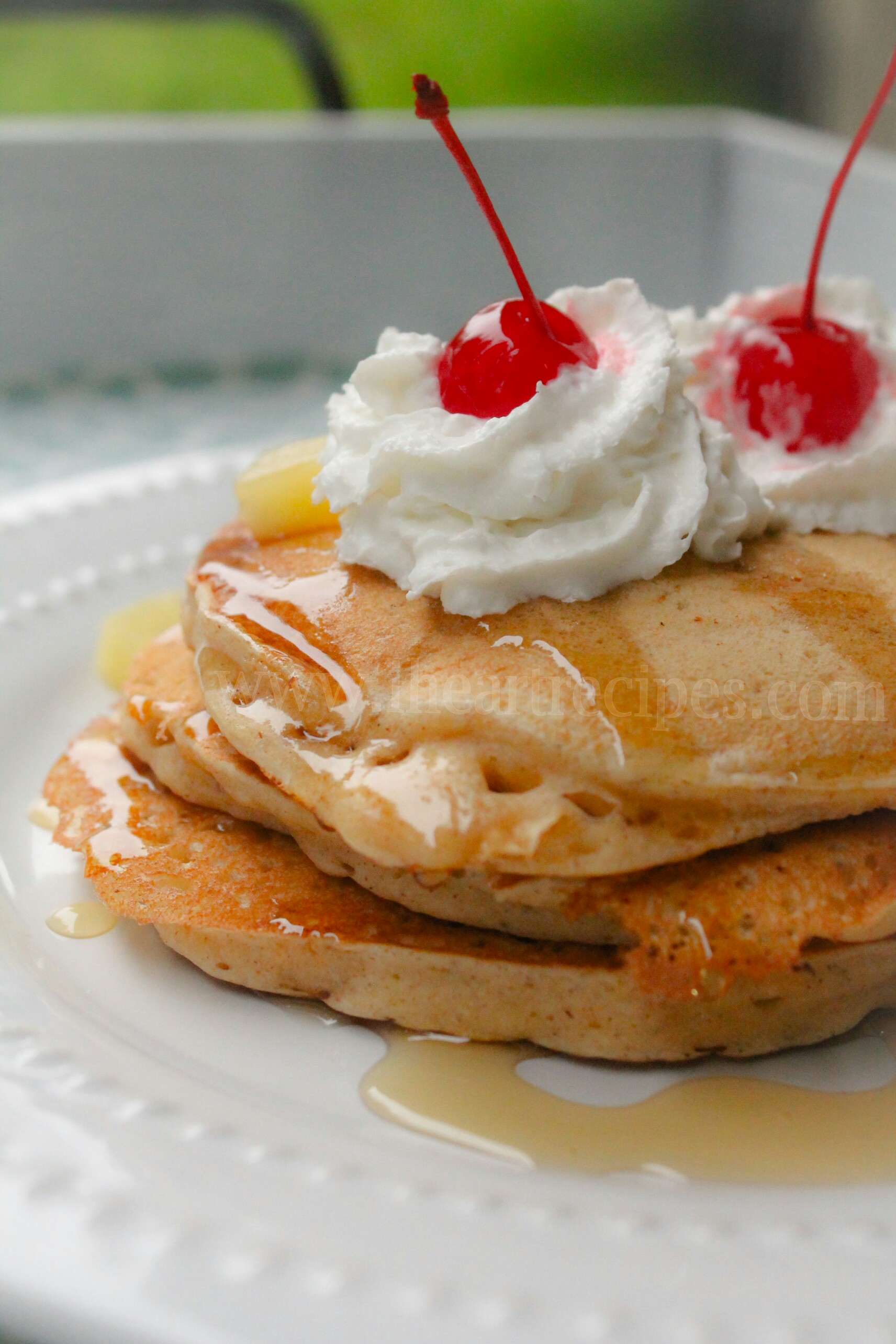 A closeup image of three pineapple pancakes topped with whipped cream, cherries, pineapple pieces, and a drizzle of honey.