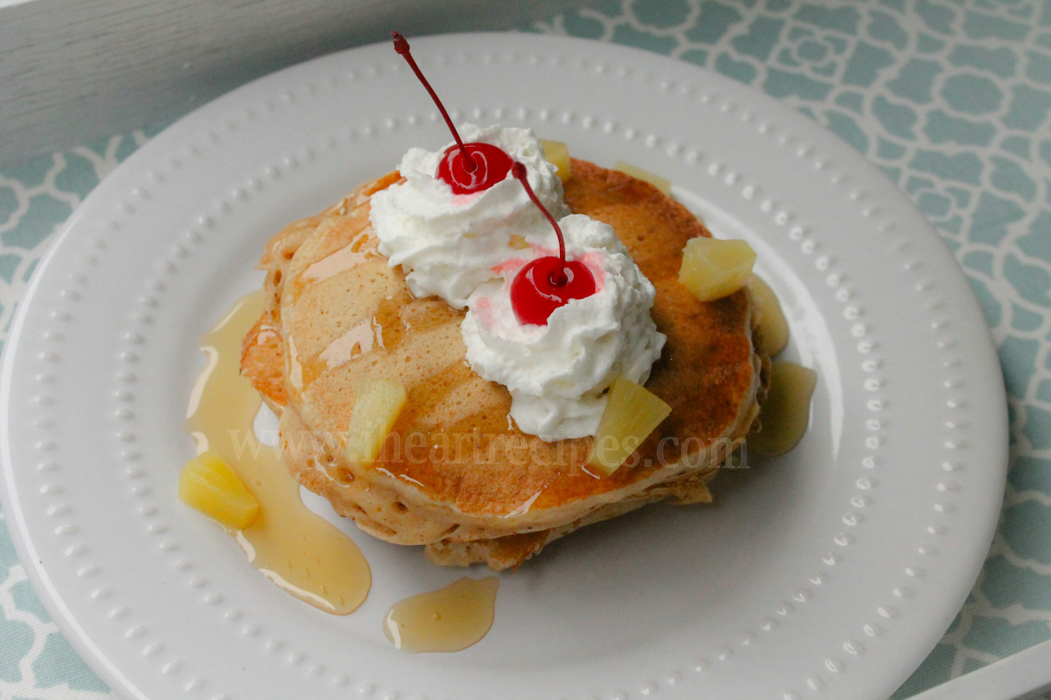 An overhead image of a stack of homemade pineapple upside down pancakes served on a white plate, topped with whipped cream, honey drizzle, cherries, and pineapple pieces.