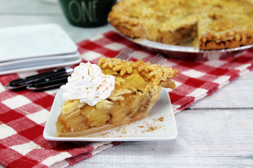 A single slice of homemade apple pie served on a small white plate, dusted with cinnamon and topped with whipped cream.
