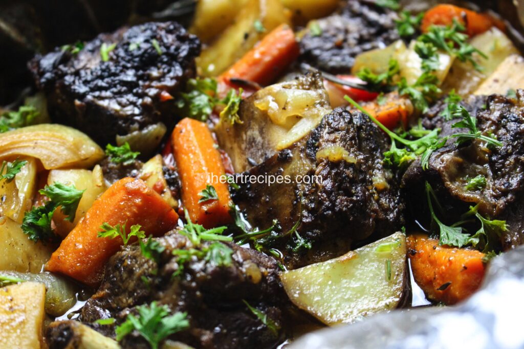 A close up of moist slow cooker short ribs alongside tender potatoes, carrots and onions garnished with bright green parsley.