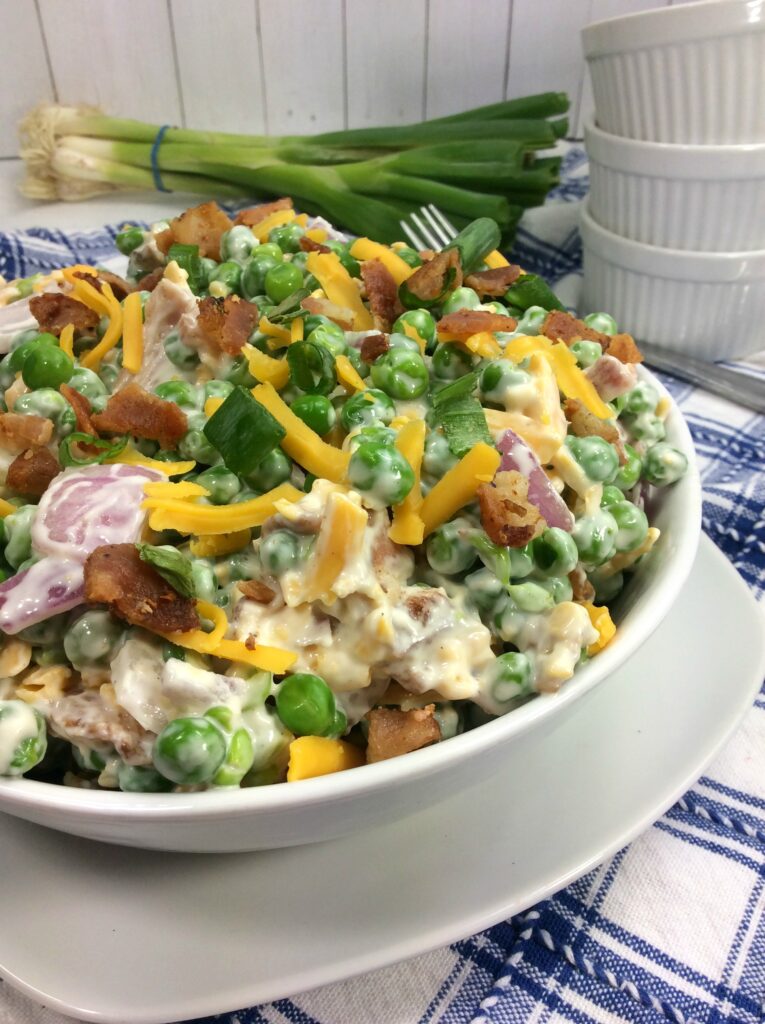 A round white bowl is filled with creamy, crunchy and colorful Simple Pea, Bacon and Cheddar Salad. 