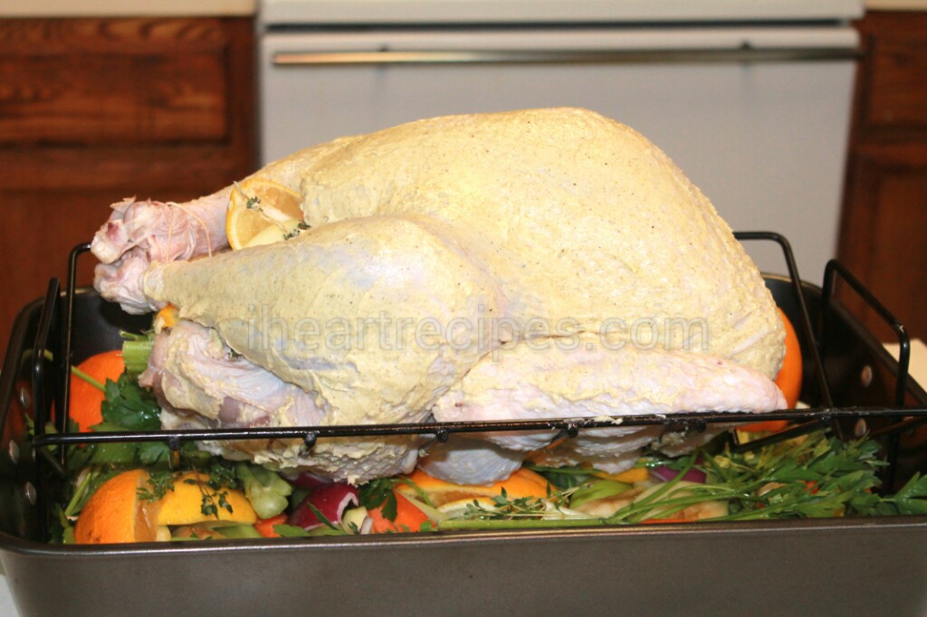 A whole raw, prepped turkey sits on a roasting pan over a bed of fruits, vegetables, and herbs. The skin of the turkey is covered in an herb and butter mixture.