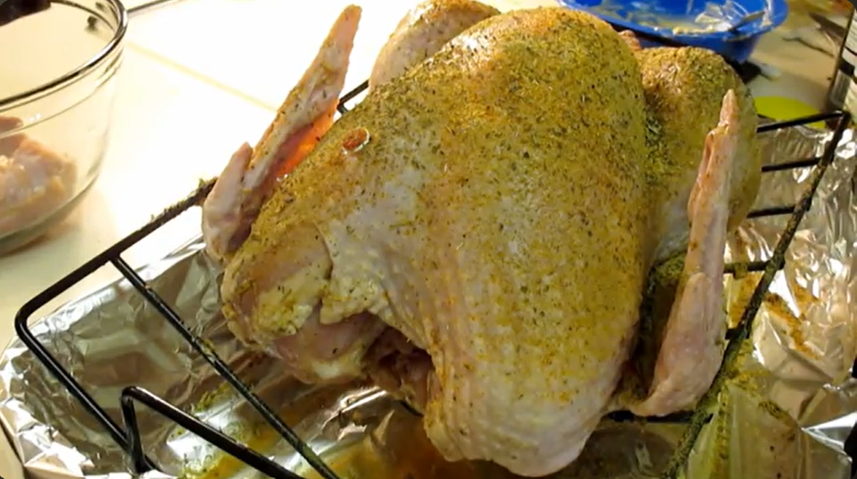 A raw turkey rests in a roasting pan, covered with a mixture of savory seasonings and spices.